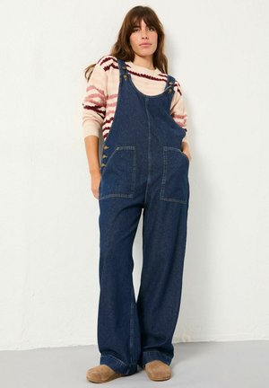 Woman with brown hair wearing a striped cream and maroon sweater, dark blue denim overalls, and tan slip-on shoes, standing against a white wall.