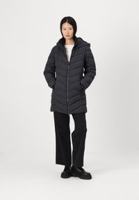 Zwarte pufferjas met capuchon, heeft een chevron-quiltpatroon, een ritssluiting en zijzakken. G gedragen over een lichte top en donkere broek.