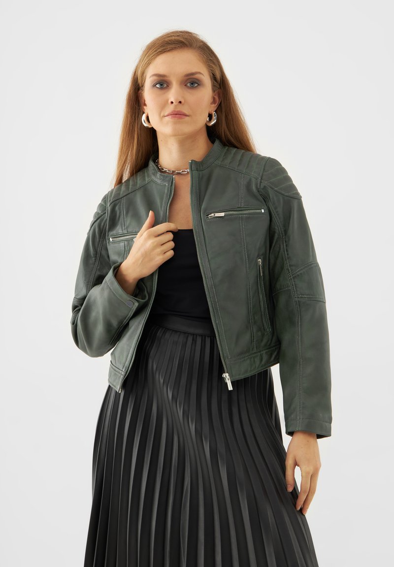 Tummanvihreä nahkatakki, jossa on pyöreä kaulus, vetoketjulliset taskut ja koristeompeleet. Yhdistetty mustan laskostetun hameen kanssa.