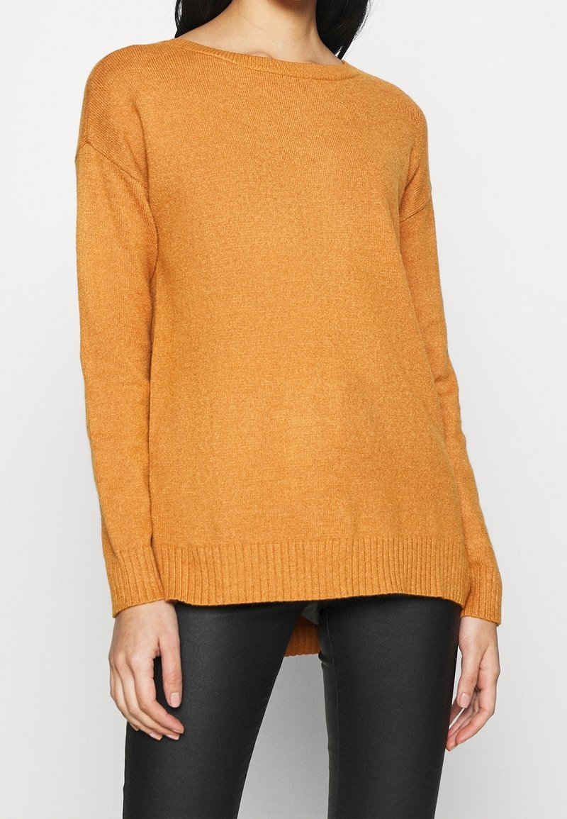 Femme portant un pull orange ample à manches longues avec poignets et ourlet côtelés, associé à un pantalon noir ajusté, debout devant un fond blanc.