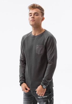 Dunkelgraues Langarmshirt mit strukturiertem Muster, ausgestattet mit einer Brusttasche und kontrastierenden Raglanärmeln. Kombiniert mit abgewetzten Jeans.