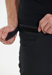 Pantalons noirs avec une texture lisse et une fermeture réglable par Velcro à la taille. Comprend des poches latérales et des détails cousus.