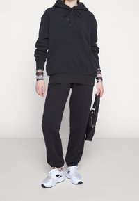 Černá fleecová mikina s kapucí, nastavitelnými šňůrkami a klokankou, v kombinaci s černými zúženými kalhotami. Obuv tvoří bílé a černé sportovní tenisky.