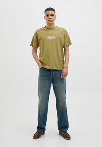 Olīvu zaļš t-krekls ar apaļu kaklu un īsām piedurknēm, ar baltu "Jack & Jones" logotipa druku. Saskaņots ar ziliem džinsu biksēm un brūnām apaviem.