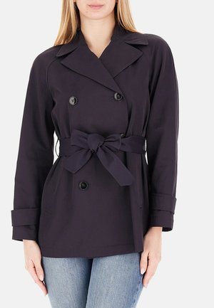 Donna che indossa una giacca doppiopetto blu scuro con cintura, ampi rever e maniche risvoltate, abbinata a jeans azzurri.