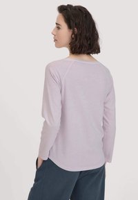 Helllila Langarmshirt, glattes Material, abgerundeter Saum und Raglanärmel. Modell von hinten betrachtet, kombiniert mit dunklen Hosen.
