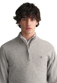 Grauer Reißverschluss-Pullover aus weichem Stoff, mit einem gerippten Kragen und kleinem gesticktem Logo auf der Brust. Glatte Textur, lange Ärmel.