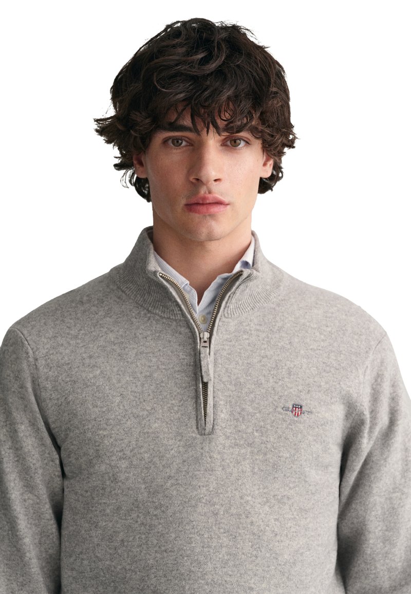 Grauer Reißverschluss-Pullover aus weichem Stoff, mit einem gerippten Kragen und kleinem gesticktem Logo auf der Brust. Glatte Textur, lange Ärmel.