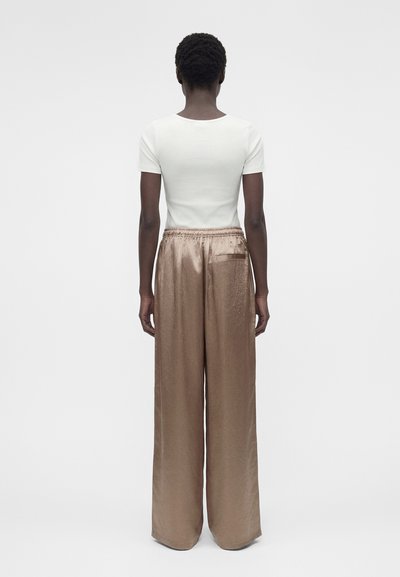 Άτομο στέκεται με την πλάτη γυρισμένη φόρεμα στενό λευκό κοντομάνικο τοπ και φαρδιά, γυαλιστερά μπεζ παντελόνια με ελαστική μέση.