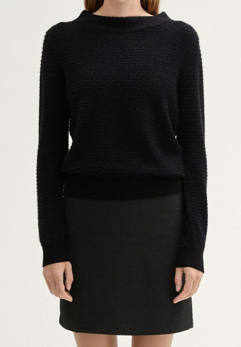 Femme portant un haut noir texturé à manches longues et une jupe noire assortie, debout devant un fond clair uni.