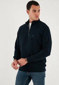 Sötétkék, cipzáras kapucnis pulóver, amely puha anyagból készült, álló gallérral, bordázott mandzsettákkal és egy kis logórésszel a mellkasán.