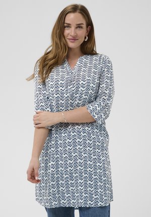 Kobieta ubrana w tunikę w biało-granatowy zygzakowaty wzór z rękawami do łokcia, niebieskie dżinsy, srebrne kolczyki koła i srebrna bransoletka.