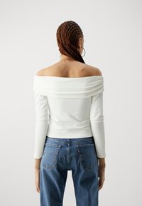Weißes, schulterfreies Oberteil mit langen Ärmeln und gerafftem Stoffdetail. Kombiniert mit blauen Jeans aus Denim im klassischen Fünf-Taschen-Design.