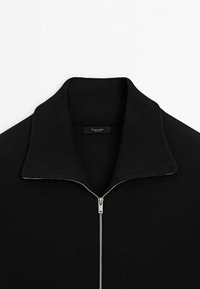 Pull en tricot noir avec une fermeture éclair argentée partiellement ouverte et un large col côtelé, montré sur un fond blanc.