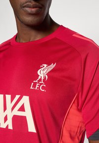Red athletic shirt with a round neckline. Features a white Liverpool FC logo and a contrasting patterned texture on the side.