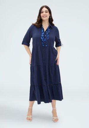 Kobieta uśmiechająca się, ubrana w granatową sukienkę midi zapinaną na guziki z podwiniętymi rękawami, niebieski efektowny naszyjnik i białe sandały na paski, stoi z rękami w kieszeniach.