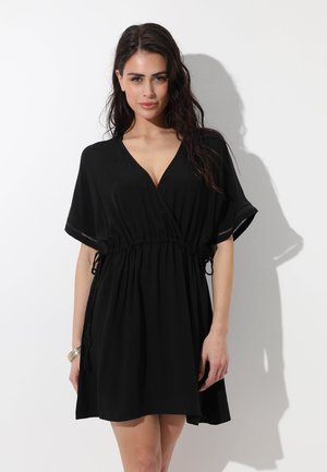 Donna con capelli lunghi e scuri che indossa un vestito nero corto con vita stretta e maniche corte, in piedi davanti a uno sfondo bianco.