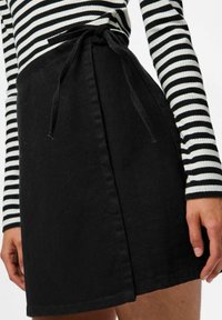 Jupe portefeuille noire avec ceinture nouée portée avec un haut à manches longues noir et blanc rayé, montrant les mains sur les côtés.