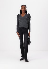 Black puff-sleeve, sparkly V-neck top paired with fitted black jeans. Model holds a small black bag. Footwear is pointed black heels.