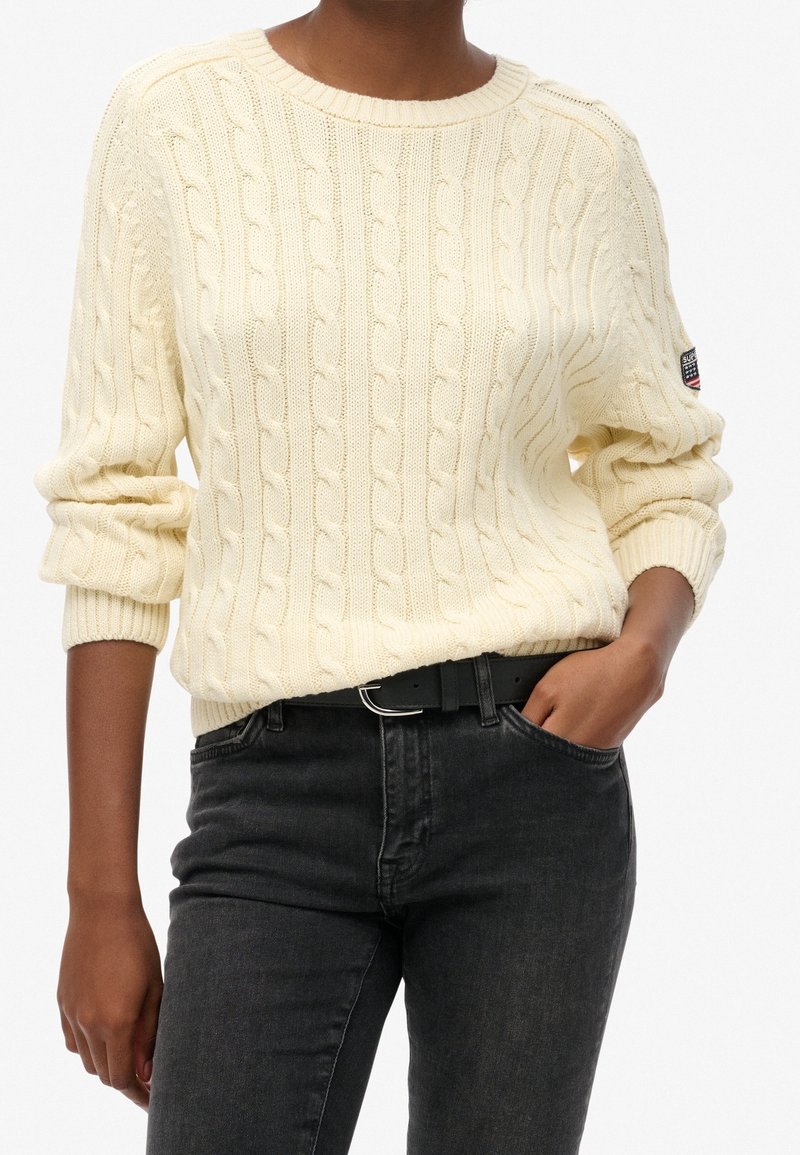 Cream cable-knit sweater with a crew neckline and rolled sleeves, paired with dark denim jeans and a black belt. Features a small logo patch.
