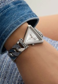 Reloj triangular plateado con esfera blanca, marcada "GUESS," que presenta una pulsera de eslabones con acentos plateados y oscuros alternos.