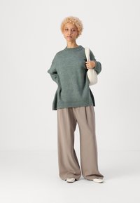 Maglione verde oversize in maglia con polsini a coste, abbinato a pantaloni beige larghi. La modella tiene una borsa bianca, in piedi su uno sfondo bianco.