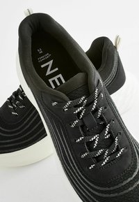 Black athletic sneakers featuring a textured fabric upper with white striped accents, round laces, and a cushioned white sole for comfort.