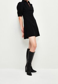 Vestido negro con cuello, mangas abullonadas y dobladillo acampanado. Combinado con botas negras de punta fina hasta la rodilla. Tela suave, diseño minimalista.