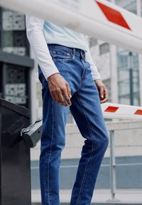 Jeans de mezclilla en azul oscuro, de corte ajustado. Presenta bolsillos estándar, textura desgastada y costuras en contraste. Combinados con una camisa de manga larga clara.