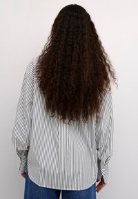 Czarny i biały pasiasty longsleeve, charakteryzujący się luźnym krojem, zaokrąglonym dołem i mankietami zapinanymi na guziki. W zestawie z wysokim stanem niebieskimi dżinsami.