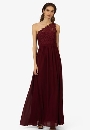 Abito bordeaux monospalla con corpetto in pizzo floreale, gonna in chiffon fluente e vita arricciata. Sandali completano l'aspetto elegante.