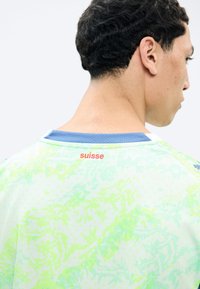 Junger Mann mit kurzen lockigen Haaren, der sich von der Kamera abwendet, trägt ein hellgrünes und blaues Hemd mit dem Schriftzug "suisse" in Rot auf dem oberen Rücken.