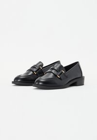 Schwarze Lederloafers mit niedrigem Blockabsatz, die über ein genähtes Design und goldfarbene Hardware verfügen, die den vorderen Riemen akzentuiert.