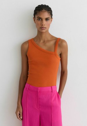 Vrouw met gevlochten haar draagt een asymmetrisch oranje hemdje en felroze broek met hoge taille, staand tegen een witte muur.