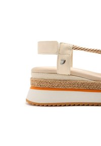 Sandalia de plataforma color beige con una suela intermedia de yute trenzado, tira de acento naranja, suela de goma texturizada y correa ajustable. Detalle del logo en plata visible.