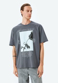 Camiseta de algodón gris con cuello redondo, que presenta una impresión gráfica de una rosa y texto debajo. Manga corta, corte holgado, diseño casual.