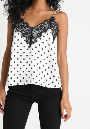 Femme portant un haut blanc sans manches à pois noirs avec un bord en dentelle noire, associé à un pantalon noir, sur un fond uni.