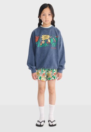 Niña joven con trenzas, vistiendo un suéter azul con flores, pantalones cortos con estampado, calcetines blancos y sandalias negras, de pie frente a un fondo liso.