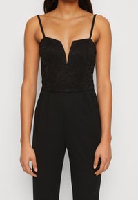 Mono negro con parte superior de encaje y escote en V profundo, tirantes finos y cintura ajustada que lleva a pantalones de pierna ancha.
