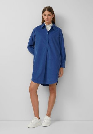Abito camicia blu, a maniche lunghe, con colletto, placket frontale e orlo curvo. Realizzato in tessuto morbido, abbinato a sneaker bianche.