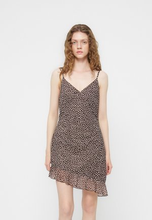 Jeune femme aux cheveux ondulés porte une mini robe marron sans manches à pois blancs avec un ourlet volanté sur un fond uni.