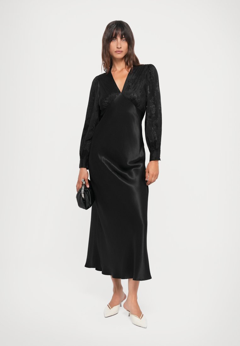 Robe longue en satin noir avec de longues manches en dentelle, décolleté en V et jupe fluide. Portée avec des chaussures blanches et un petit sac à main noir.