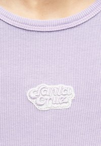 Κοντινό πλάνο ανοιχτόμωβ ριμπ ύφανσης κορυφής με λευκό κεντημένο έμβλημα «Santa Cruz» κοντά στη γραμμή του λαιμού.