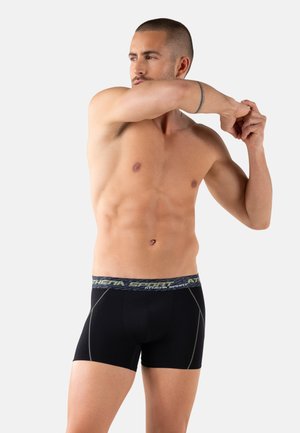 Homme musclé étirant son bras à travers sa poitrine, portant un boxer de sport noir avec une ceinture "Athena Sport", sur un fond clair.