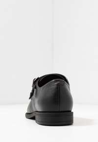 Chaussure en cuir noir avec un bout arrondi, un détail de boucle sur le côté, une texture lisse et une semelle en caoutchouc plate et basse. Vue arrière.