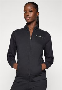 Sweat à zip noir fabriqué en tissu lisse avec un col haut, présentant un petit logo Champion blanc sur la poitrine et des poches latérales.