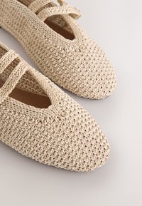 Paire de chaussures plates tissées beige à bouts fermés avec des détails de lanières sur un fond neutre.