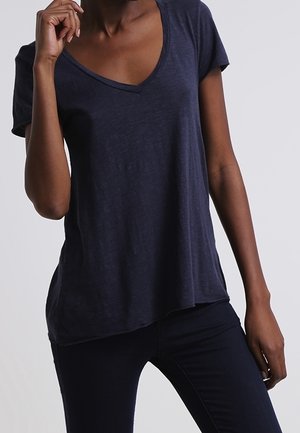 Persona vestida con una camiseta de manga corta en color azul marino liso con cuello en V y pantalones oscuros a juego, de pie con una mano cerca de su rostro.