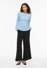 Top a maniche lunghe azzurro chiaro con finitura testurizzata e orlo smerlato, abbinato a pantaloni neri larghi e décolleté nere con punte dorate.