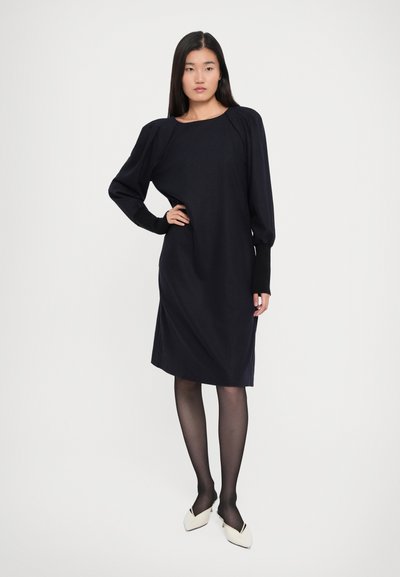 Woman stands with hand on hip wearing a black knee-length dress with puffed sleeves, sheer black tights, and white pointed shoes on white background.